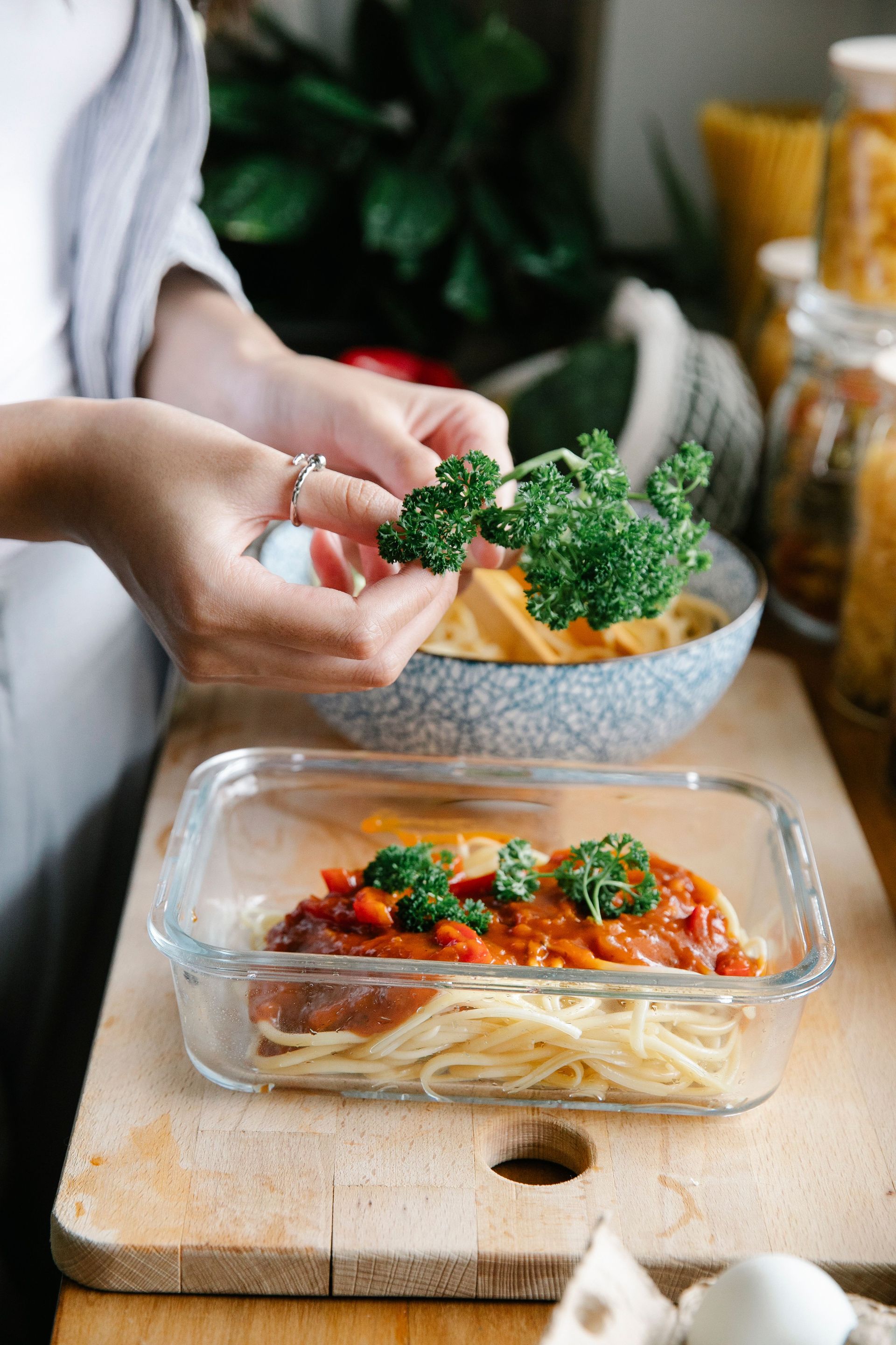 Unrecognizable female cook adding herbs into container with spaghetti and bolognese sauce while standing at counter and cooking in kitchen